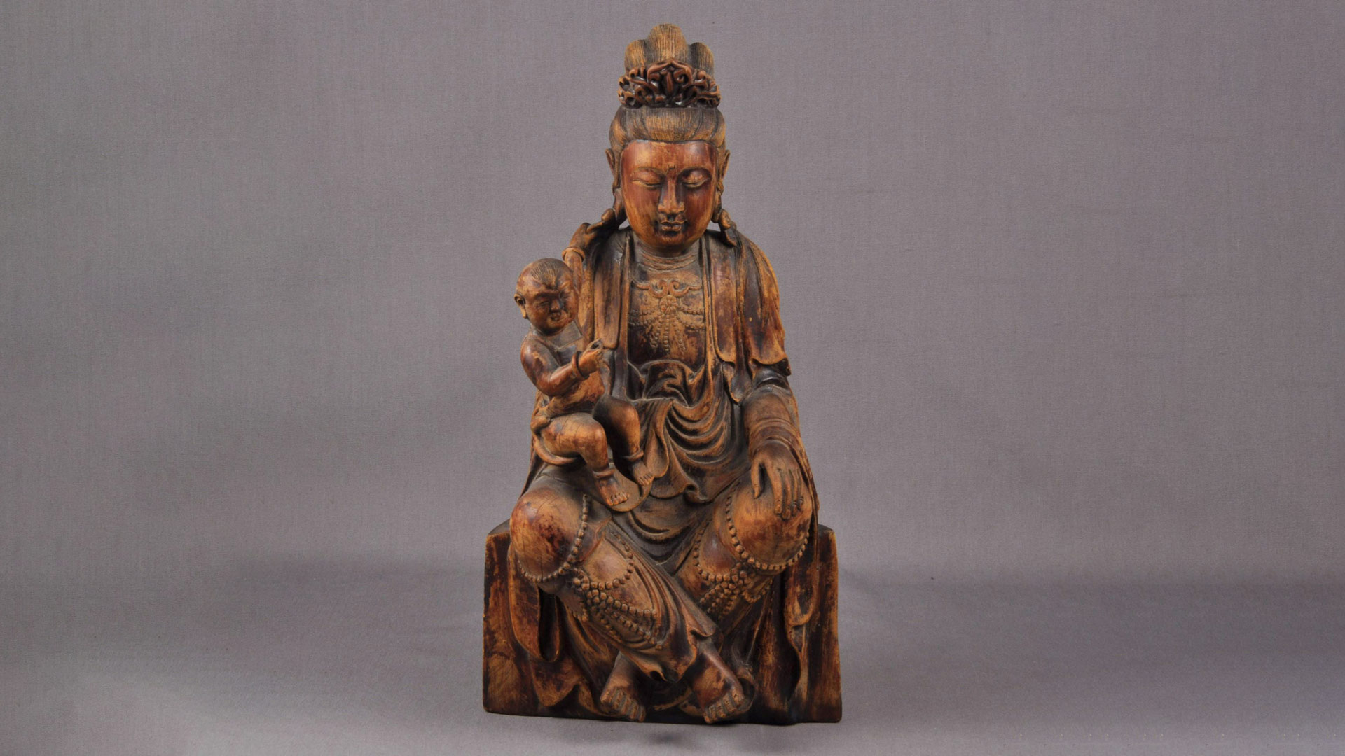 a wooden carving of a Buddhist deity sitting and dressed in a robe and various accessories while holding a child.