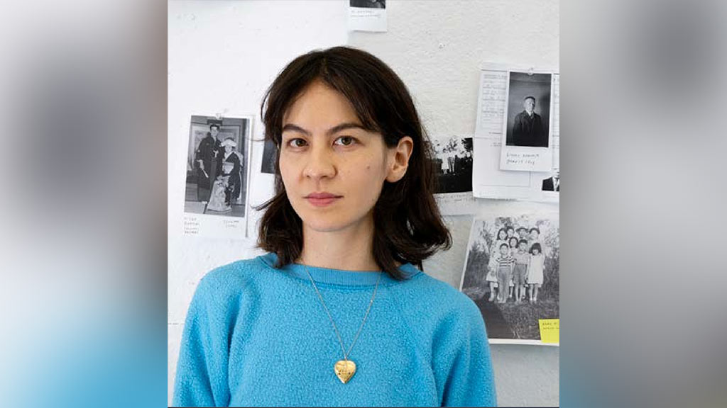 a woman with dark hair poses in front of a white wall with several black and white photographs of people.