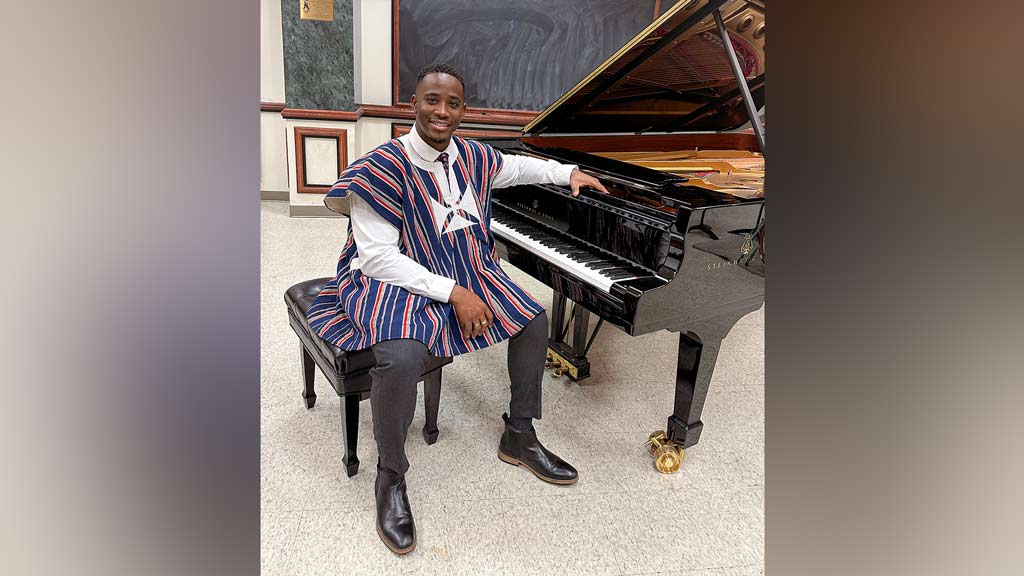 a man smiling, in vibrant, African clothing posing next to a piano.