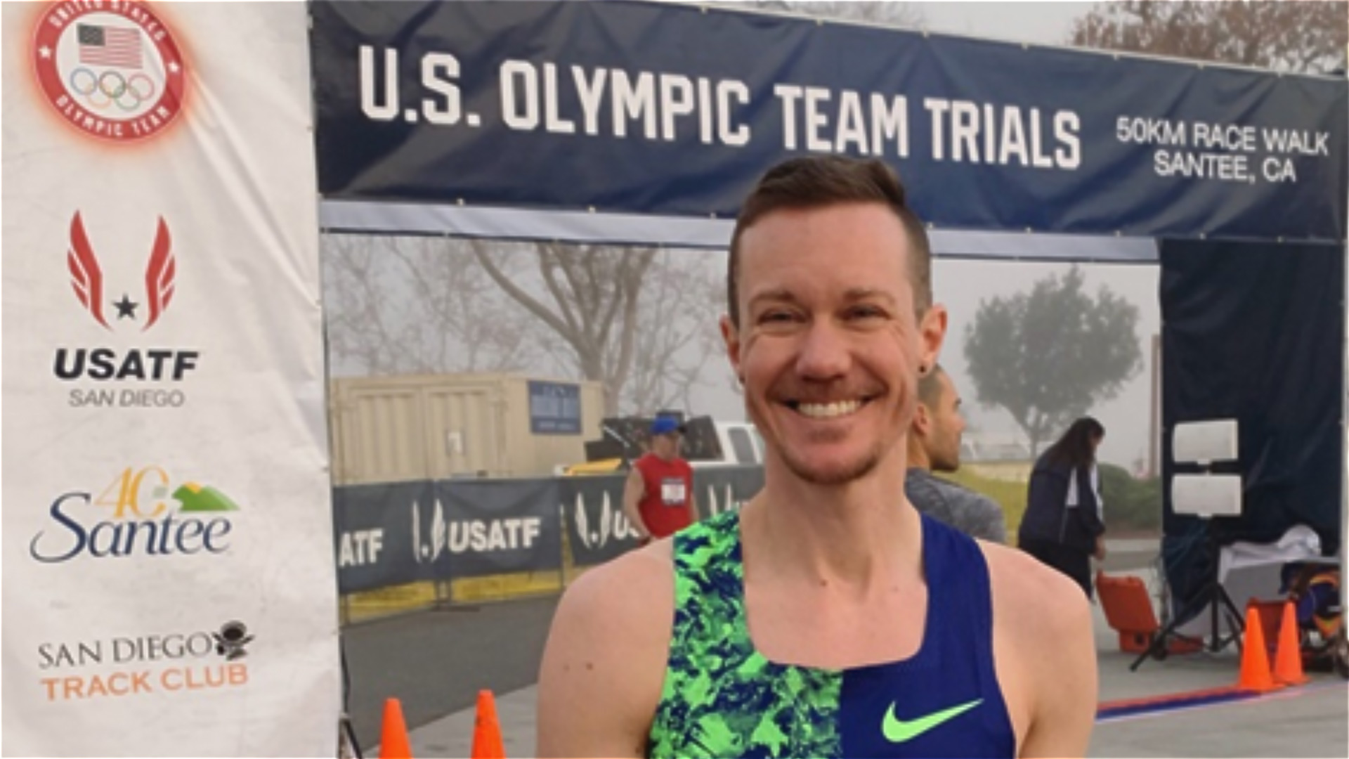 A man smiling at a racing event with a banner behind him saying 'US Olympic Team Trials'