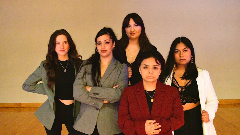 photo of 5 women dressed in blazers of various dark colors, staring at the camera with stoic, nonchalant expressions.