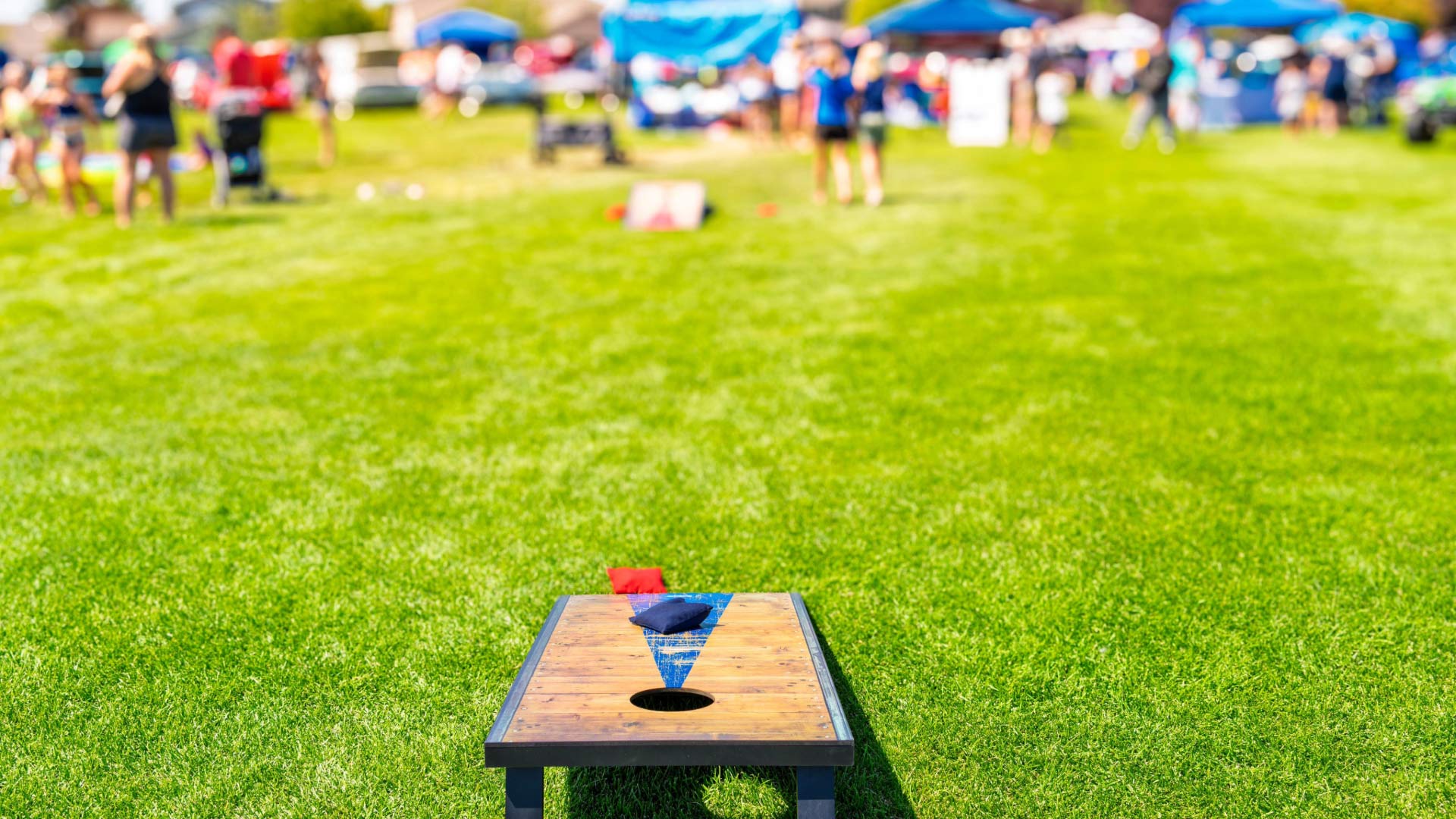 A close-up of a cornhole cover on a lawn, with a group of people and tents in the distance.