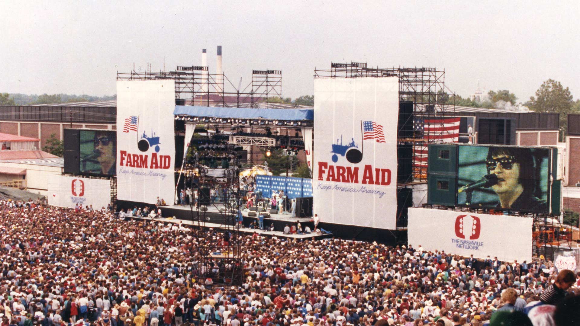 Packed outdoor concert venue with large crowd watching a performer on stage with giant banners by the stage