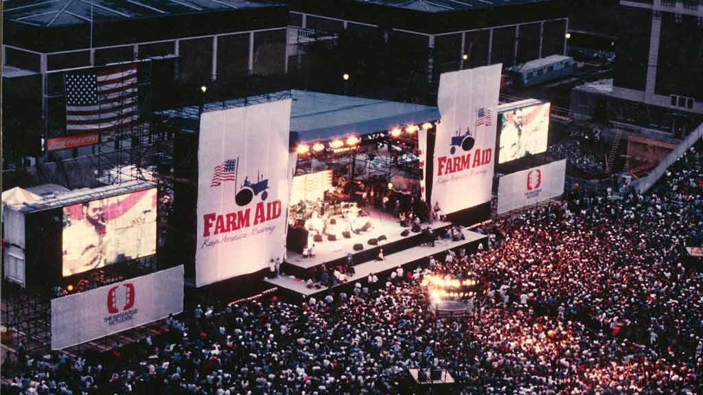 A shot of the outdoor Farm Aid festival stage and band from above, with a packed crowd in front of it.
