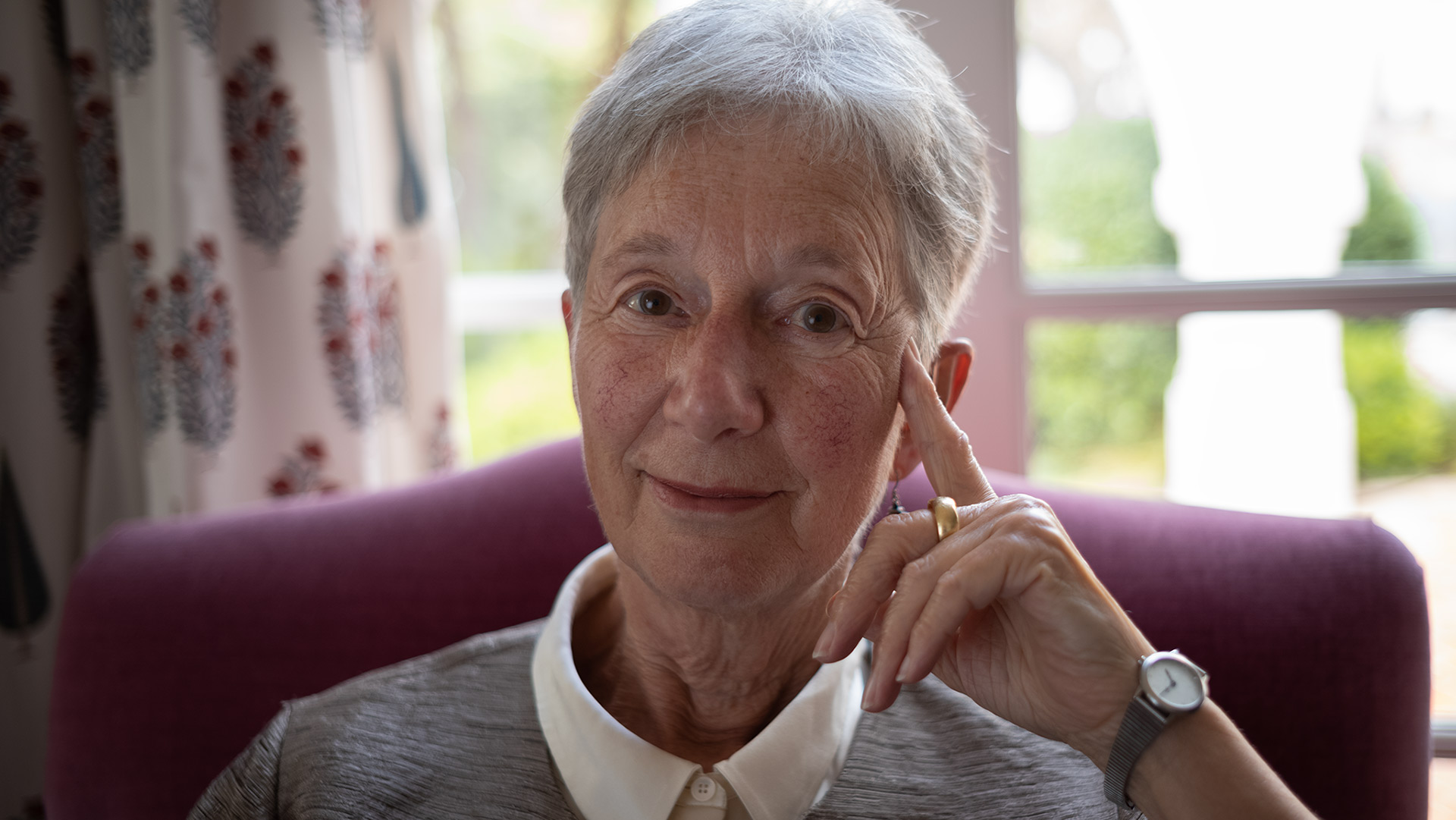 Woman sitting in a chair facing the camera in front of an window with the sky in the background