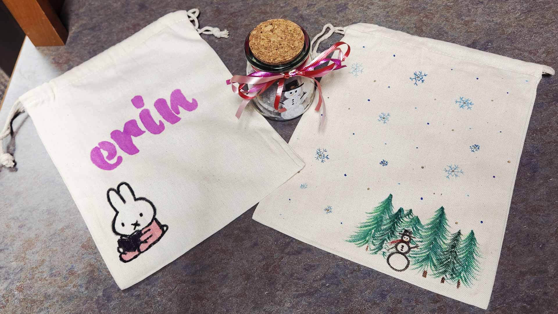 Two flat white bags and a small jar all personalized with designs, names, and winter-themed decorations