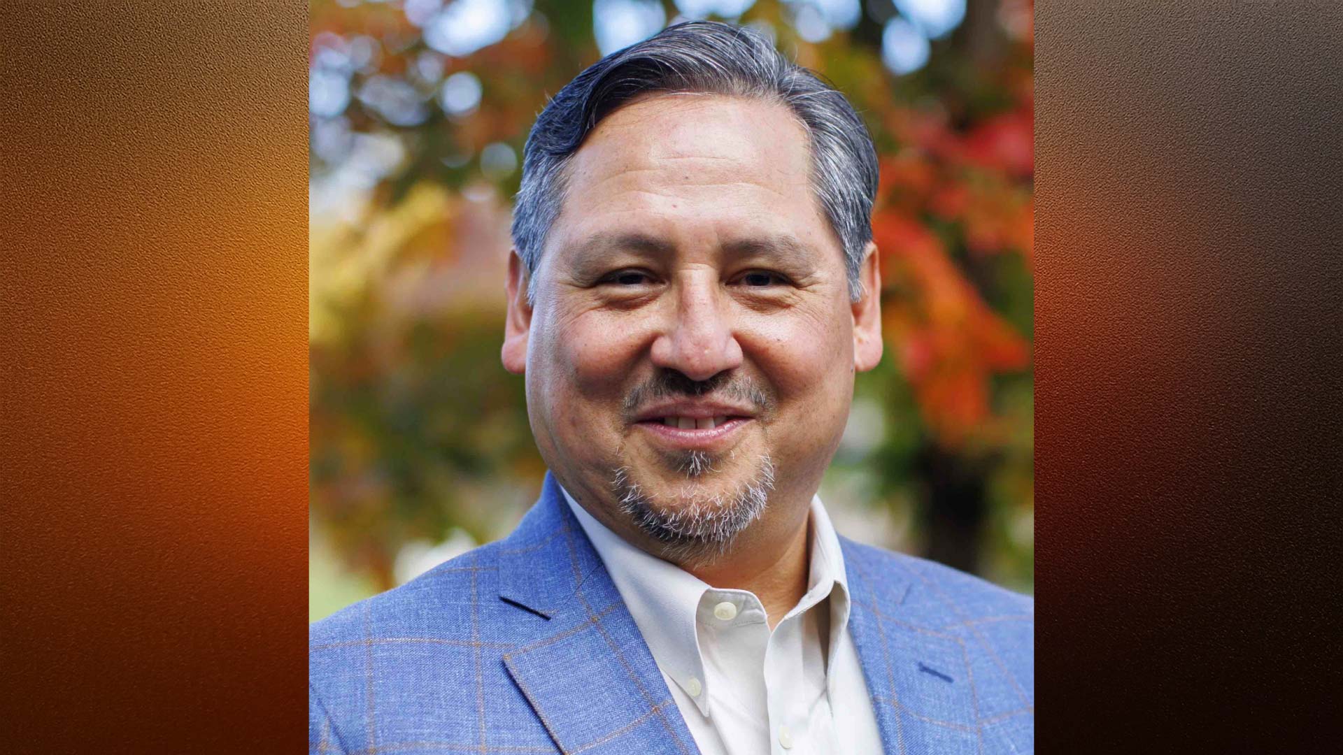 headshot of a smiling man with silvery hair and beard in front of a tree with fall colored leaves