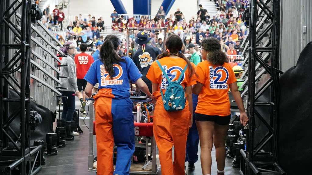 # girls in orange shirts that say "FRC 4096" walking past crowded arena stands pushing a cart.