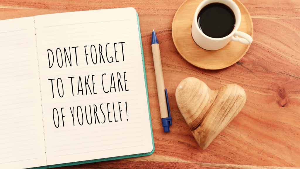 Journal placed on a wooden table reading "Don't forget to take care of yourself!". A cup of coffee, a pen, and a wooden heart lay closely to its right.