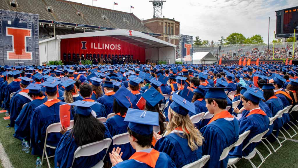 A graduation event with multiple rows of students in blue caps and gowns facing a stage