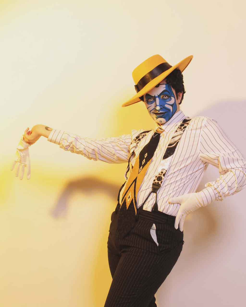 a person in white and blue face paint, tilted yellow hat, suspenders, crisp shirt and pants, and white gloves