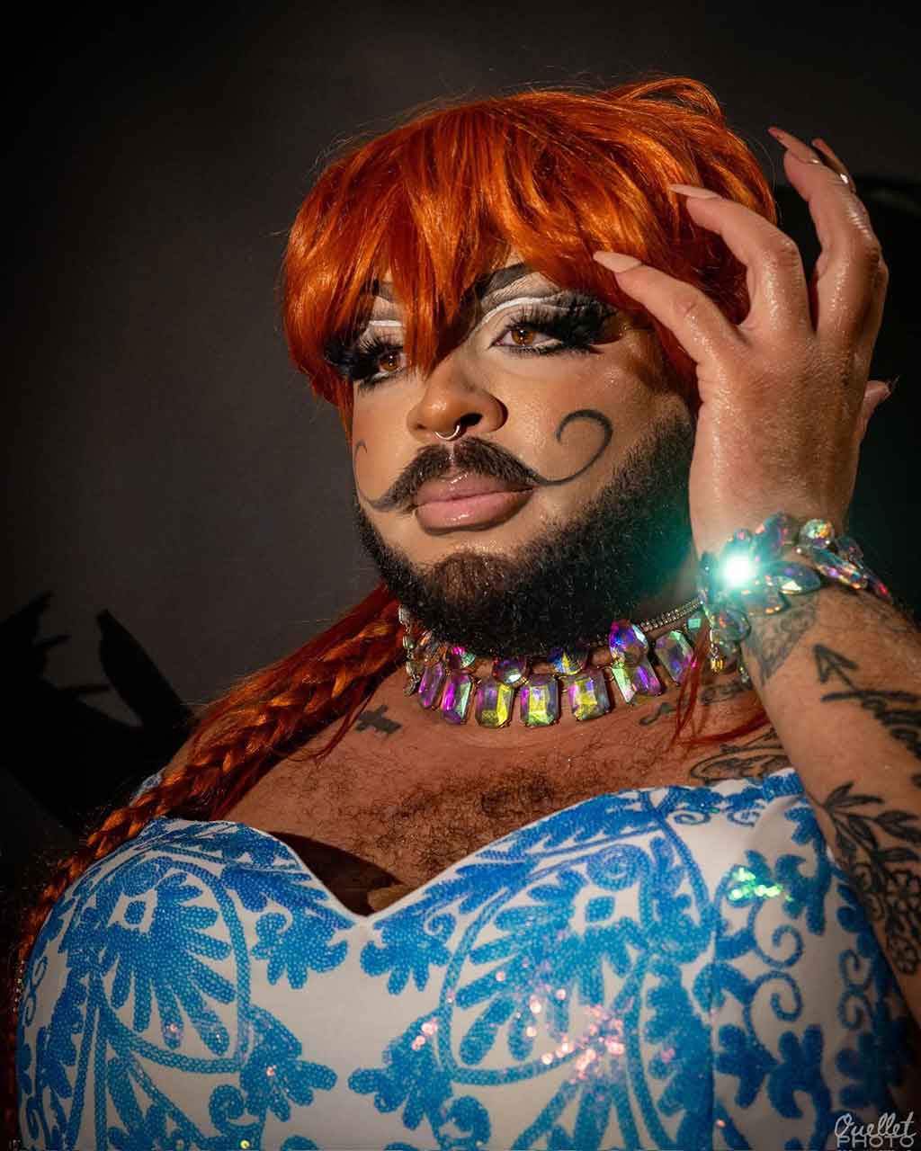 a bearded drag performer with red hair, jewels, a patterned dress, and tattoos