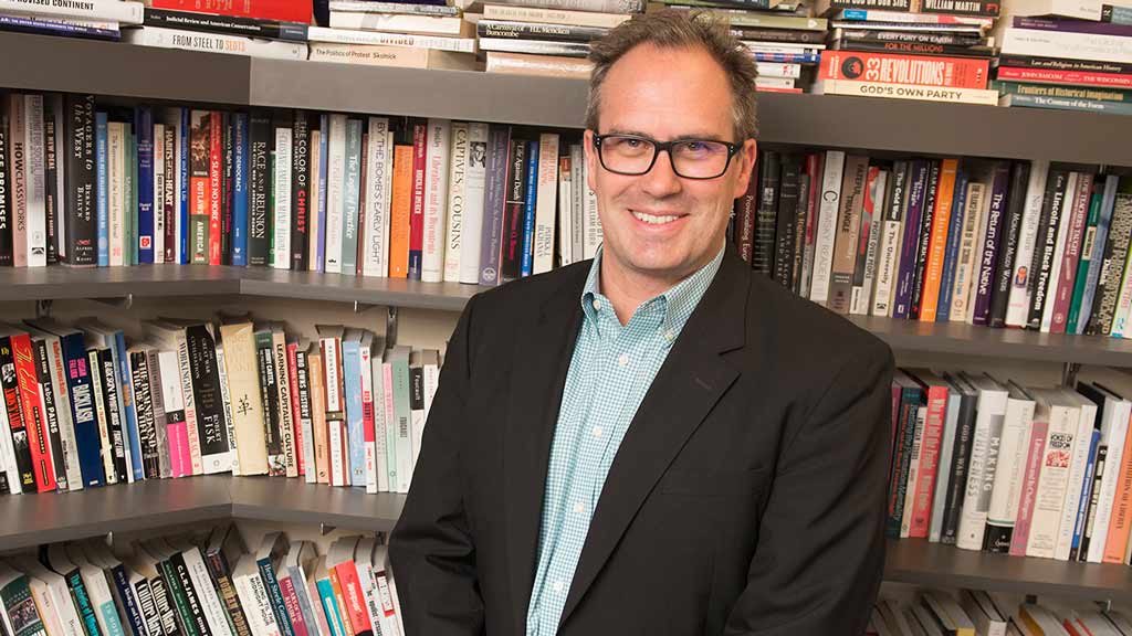 A photo of a man in a suit standing in front of full book shelves.