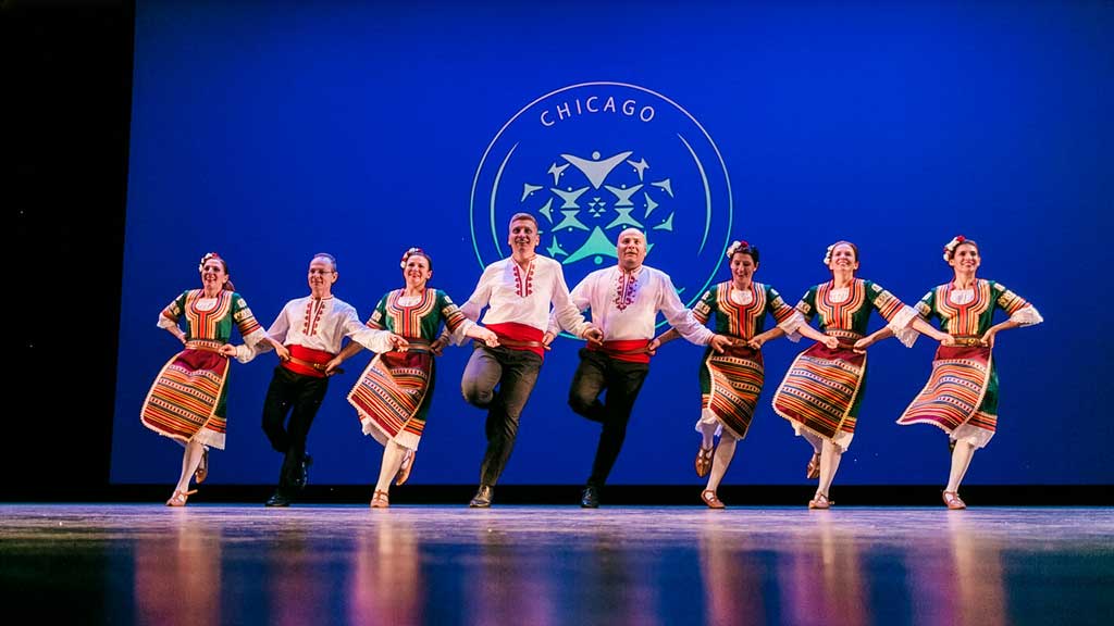 a group of approximately 8 dancers wearing traditional bulgarian costumes in a lively dance on an auditorium stage
