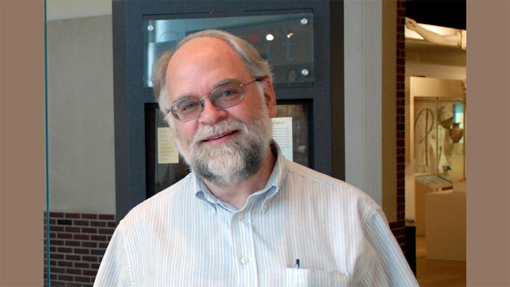 Man with beard and crisp collared shirt poses with a smile in front of an exhibit case and exhibit gallery