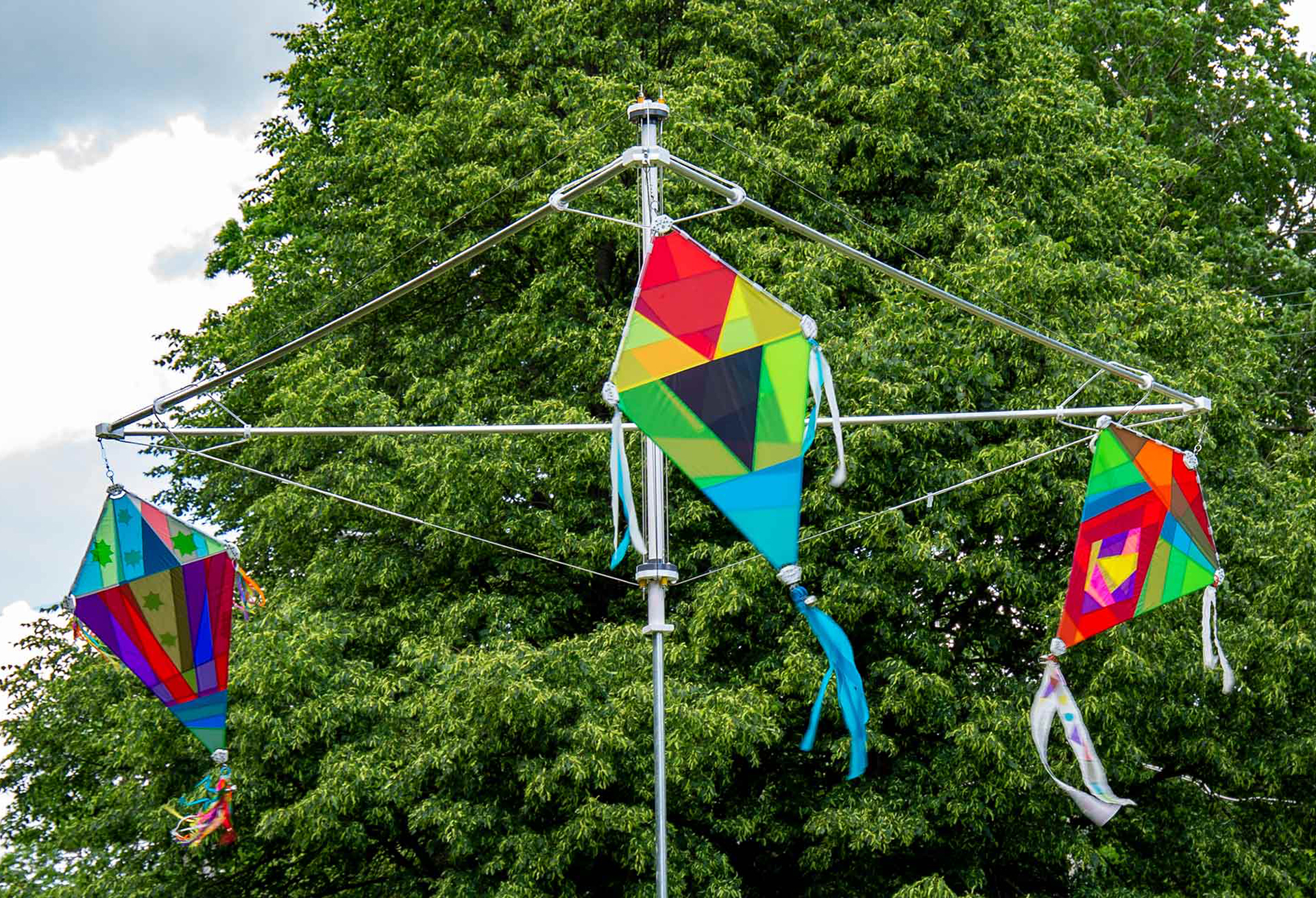 Three kites with geometric rainbow-colored patterns are hanged to a kite-shaped metal frame