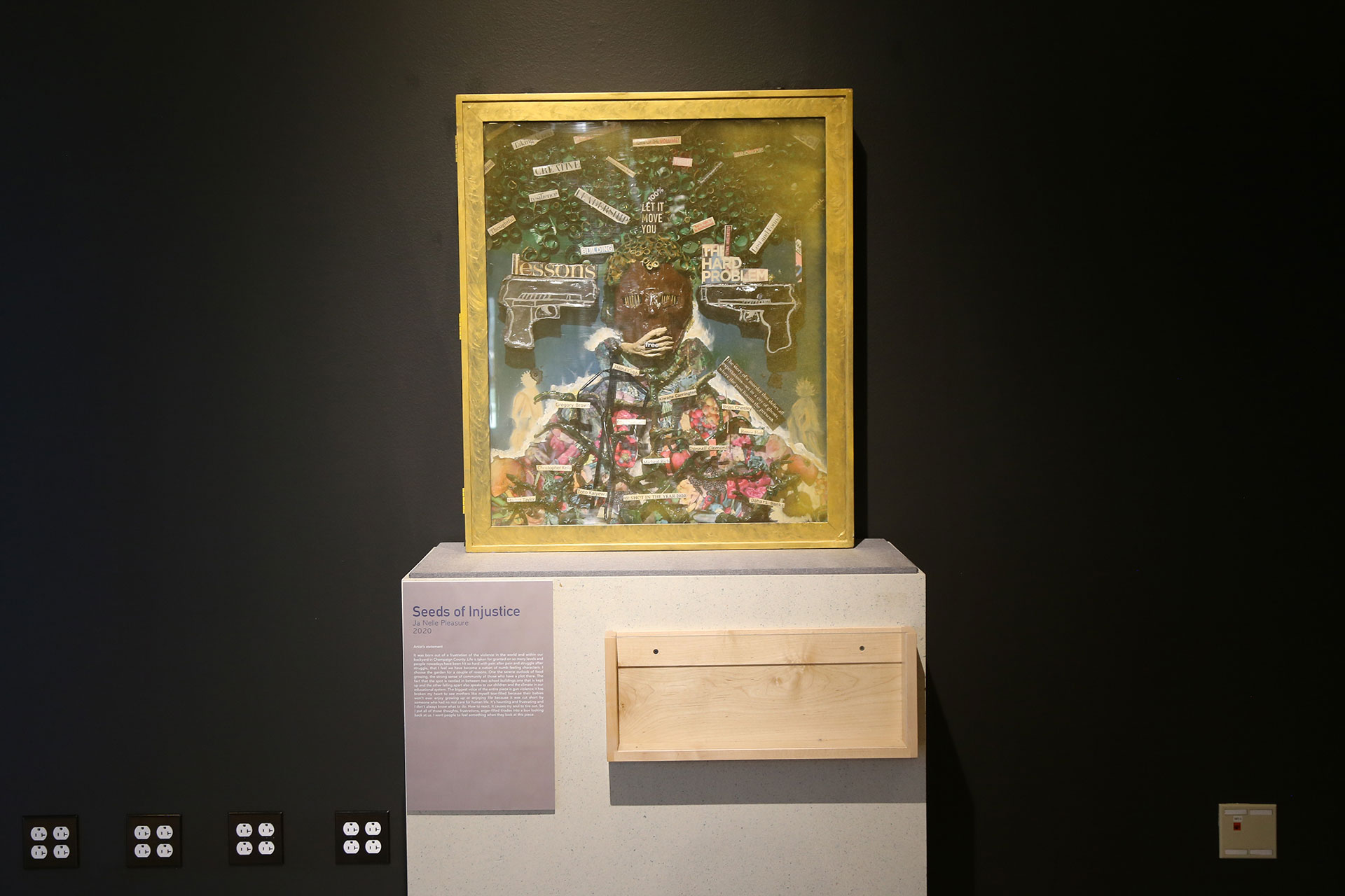 A further away look of the Seeds of Injustice with artwork placed in the middle of a white stand, the artist's statement attached to the left side and a wodden box placed on the right side
