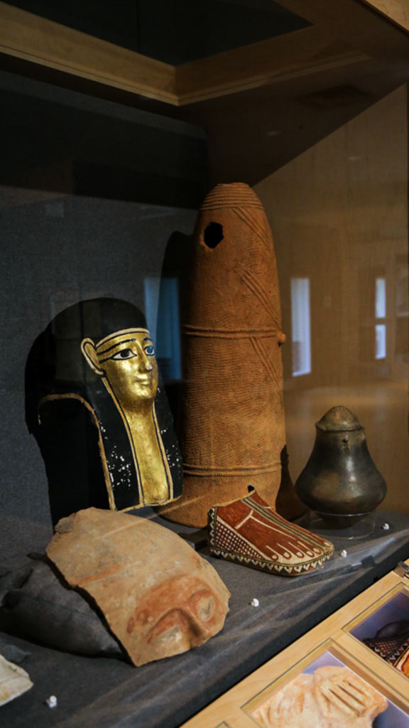Case shot with an Egyptian burial mask, an urn, a long terracotta grave marker, and fragment of a coffin lid depicting a face