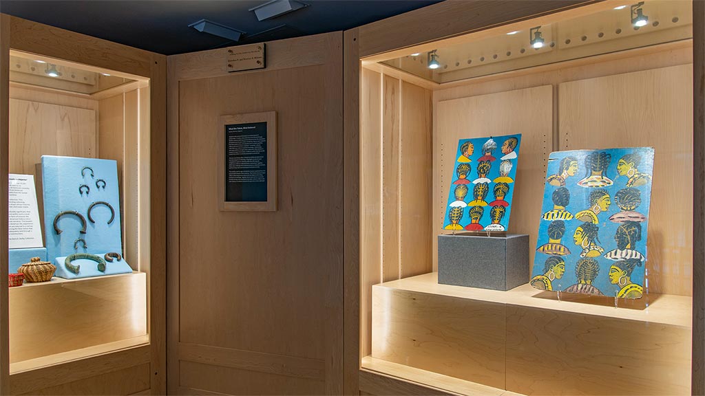 Two cases of objects featuring a blue fabric panel with round metal neck or wrist adornments and two painted signs with hairstyle diagrams