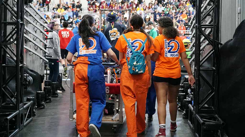 From behind, three women in oranage and blue outfits walk into a filled gymnasium