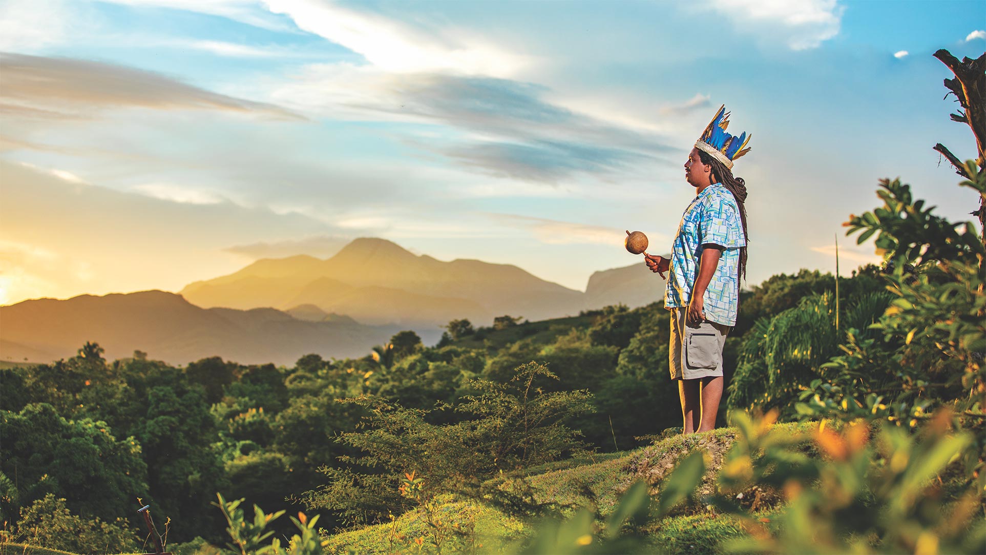 a man with feathered headwear holds a rattle type instrument outdoors in a lush mountainous landscape under a vast sky
